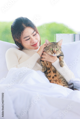 Asian young pretty female teenager girl in turtleneck sweater laying lying down on cozy comfortable white clean sheet bed under blanket smiling playing holding little cute domestic cat