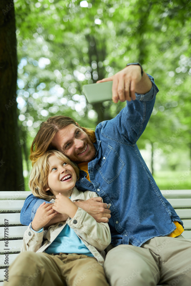 Portrait of father and son sitting on bench in park together and taking selfie photo