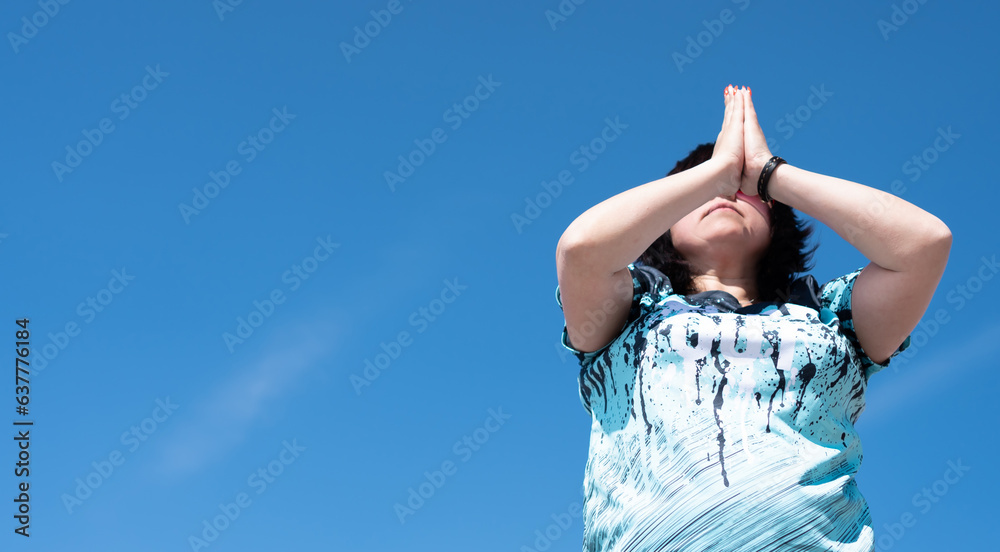 An impersonal portrait of a woman performing a set of yoga exercises, surya namaskara, holding her hands in front of her face against a blue sky.