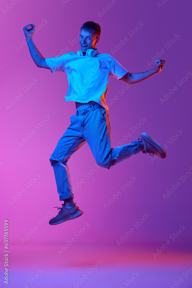 Full-length image of smiling guy in casual clothes and headphones jumping on gradient pink purple background in neon light. Excited and happy. Concept of human emotions, youth, lifestyle, fashion, ad