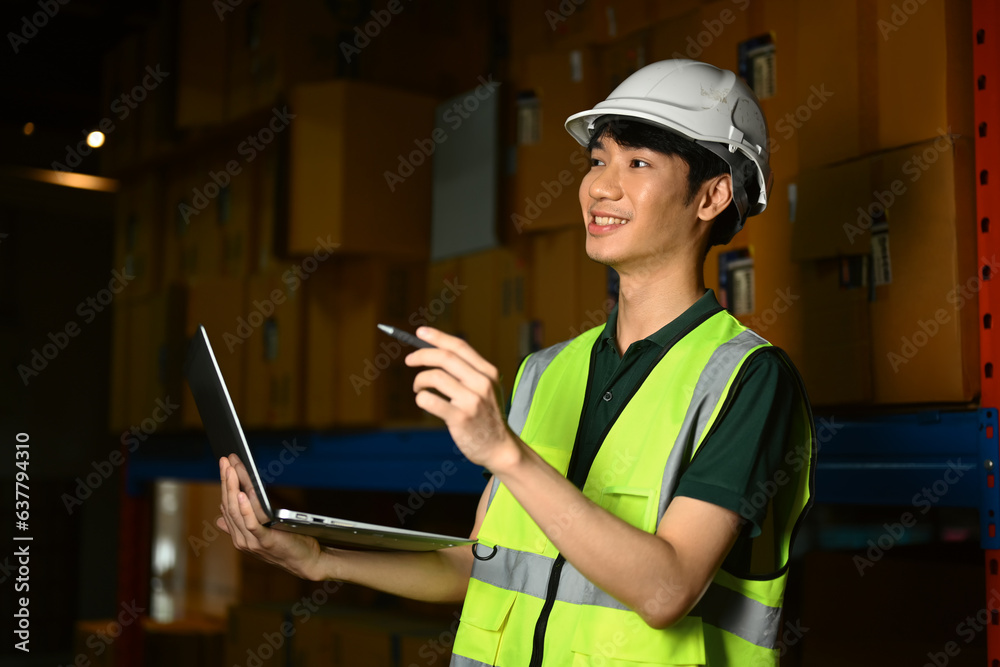 Male worker in hardhat and vest using laptop, checking inventory production stock control in warehouse Male worker in hardhat and vest using laptop, checking inventory production stock control in warehouse