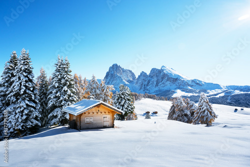 Winter landscape with wooden log cabin on meadow Alpe di Siusi on blue sky background on sunrise time. Dolomites, Italy. Snowy hills with orange larch and Sassolungo and Langkofel mountains group