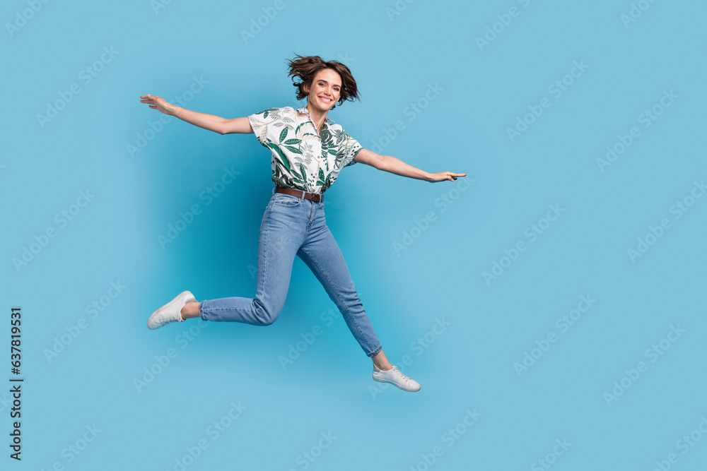 Full length photo of sweet carefree lady wear print shirt jumping high flying arms sides empty space isolated blue color background