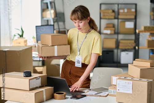 Young woman in casualwear holding stack of packed boxes and using laptop while checking personal data of customer in database photo
