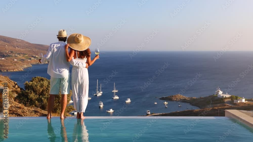A hugging, loving couple on vacation time enjoys the summer sunset over the Aegean Sea by the swimming pool with a drink