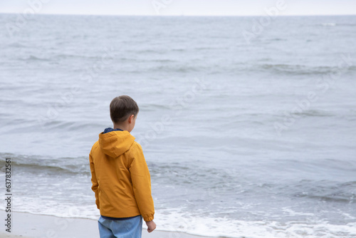A boy in an autumn yellow jacket stands on the seashore. cool autumn weather. calm sea. rear view
