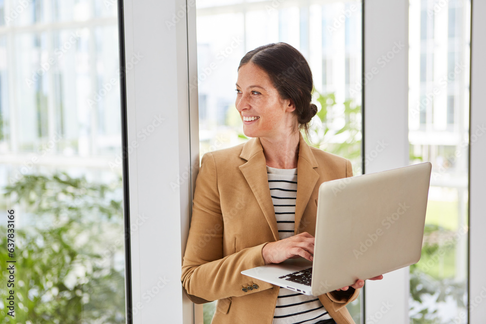 Happy businesswoman holding laptop leaning on window while day dreaming in office lounge Happy businesswoman holding laptop leaning on window while day dreaming in office lounge