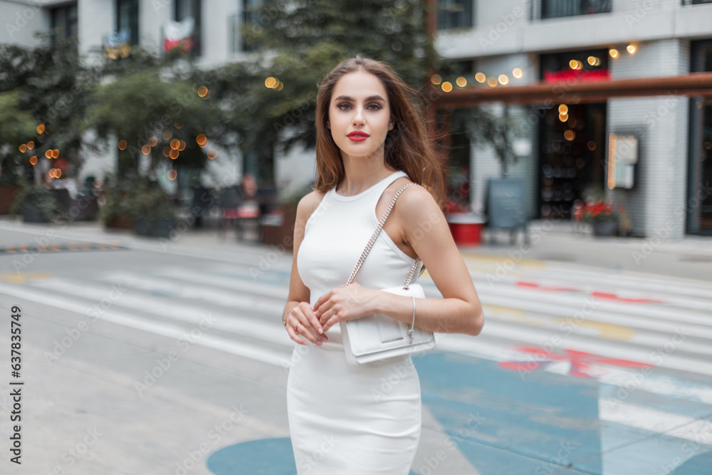 Fashionable beautiful young girl model with red lips in stylish elegant white dress with a bag walks on the street
