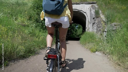 Young brown hair woman with backpack riding bicycle on park, back view. Camera moving forward after her, slow motion effect