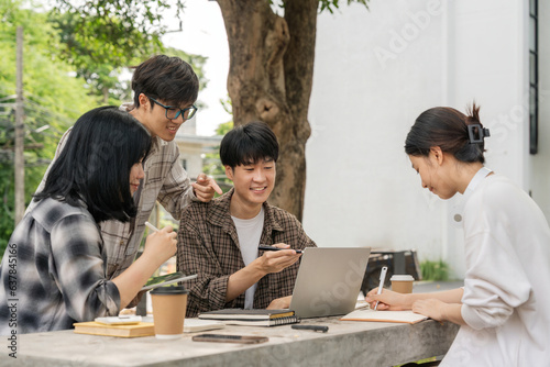 Group of happy young Asian college students sitting on a on the cement table, looking at a laptop screen, discussing and brainstorming on their university project together