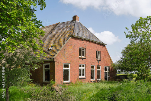 An abandoned and dilapidated farm house with red bricks and broken glass on a neglected terrain with tall grass. Concept of rural depopulation. photo