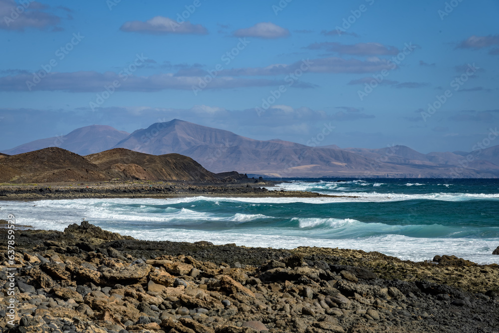 View from Lobos Island, Fuerteventura (Canary Islands). There is a rocky shore in the foreground, and a bay with blue water and breaking waves. The island of Lanzarote is visible in the background.