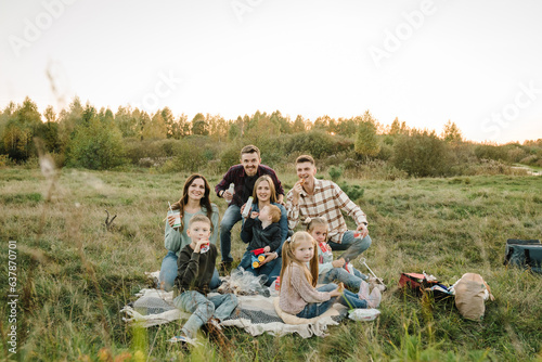 Happy big family with kids spending time together in field. Large company people eating on picnic in park. Mother, father, daughter, son sitting on blanket at sunset. Family holiday outdoors.