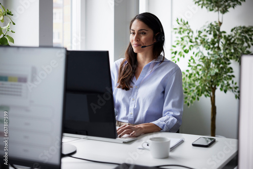 Smiling mid adult businesswoman using headset in office in front of computer screen photo