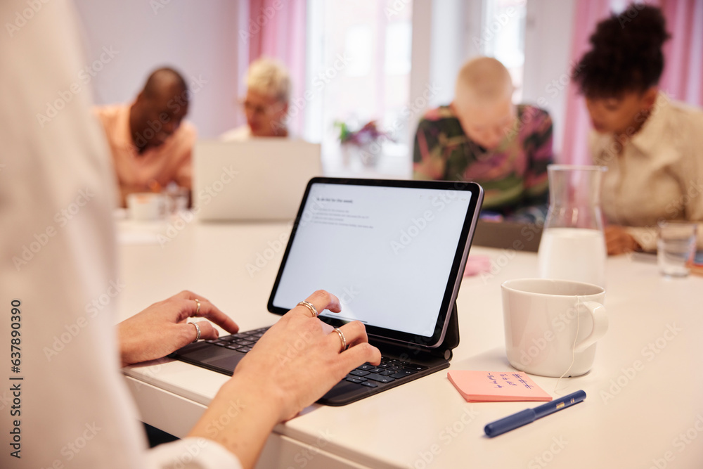 View of woman's hands using laptop, coworkers in background View of woman's hands using laptop, coworkers in background