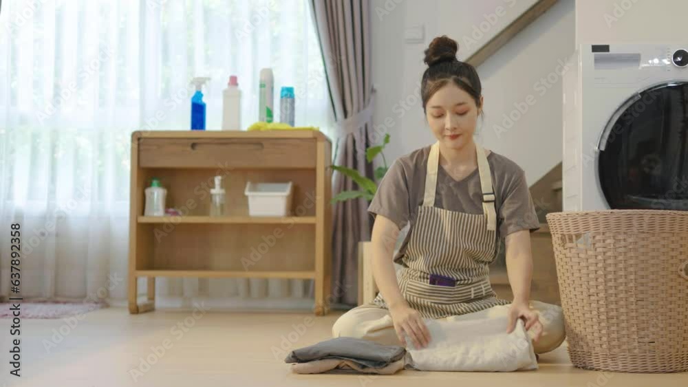Young asian woman sitting on floor folding clothes at laundry room, girl take clothes from basket and fold it and smell clothes from the wash, sort clean laundry in stack domestic chores. Young asian woman sitting on floor folding clothes at laundry room, girl take clothes from basket and fold it and smell clothes from the wash, sort clean laundry in stack domestic chores.