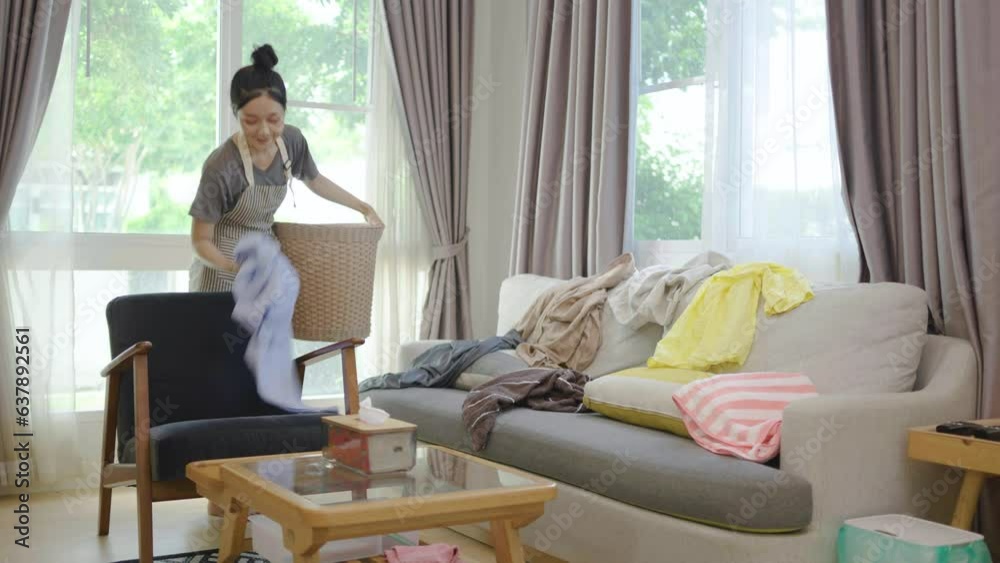 Asian woman cleaning housework in living room at home. Female young girl housekeeper take messy dirty clothes into basket on sofa for housekeeping housework chores. Asian woman cleaning housework in living room at home. Female young girl housekeeper take messy dirty clothes into basket on sofa for housekeeping housework chores.