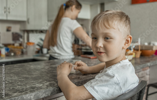 A cute blond-haired boy-child is sitting at the kitchen table, waiting for his mother to cook food and watching TV