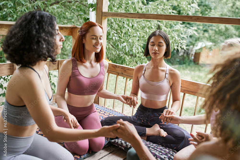 multiracial girlfriends in sportswear holding hands, sitting, meditating with closed eyes in patio multiracial girlfriends in sportswear holding hands, sitting, meditating with closed eyes in patio