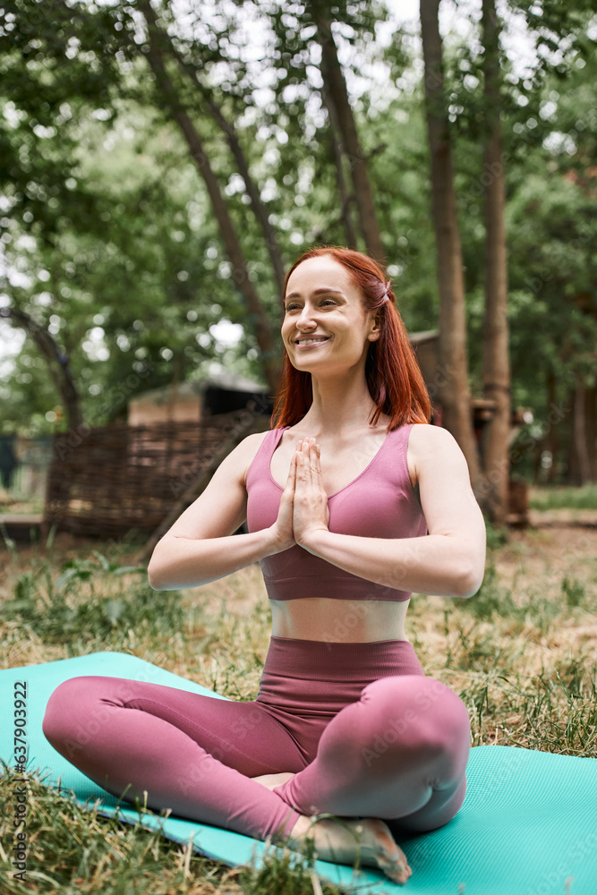 smiling redhead woman with closed eyes and praying hands meditating in park of retreat center