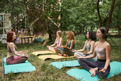 multiethnic girlfriends in sportswear meditating in lotus pose in park, inner peace, harmony