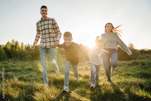 Mom, dad, daughter and son walk in green grass in spring field. Happy young family with children spending time together, running outside, go in nature at sunset. Concept of family holiday outdoors.