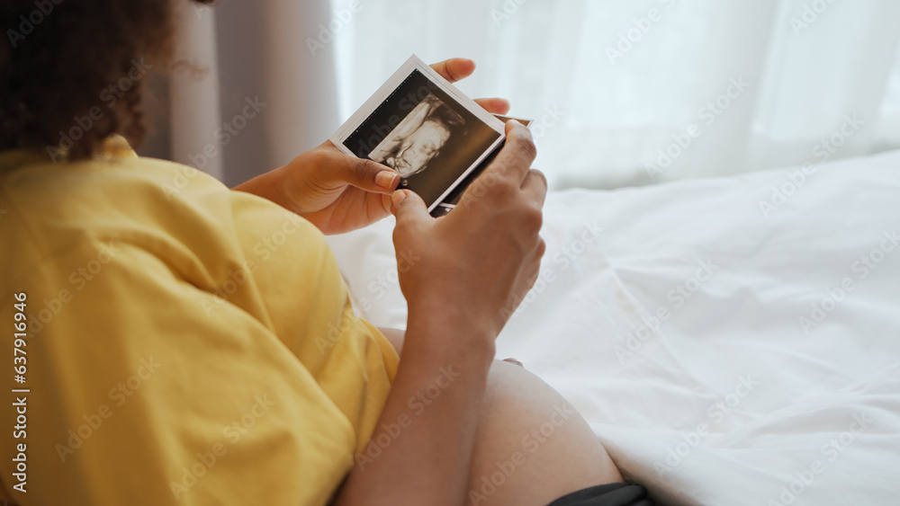 Black African American pregnant woman looking her ultrasound scan photos on bed. Expecting mother waiting her baby. Concept of pregnancy, Maternity prenatal care. Black African American pregnant woman looking her ultrasound scan photos on bed. Expecting mother waiting her baby. Concept of pregnancy, Maternity prenatal care.