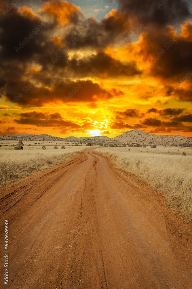 A dirt road leading off into the distance to mountains on the horizon at sunset with a dramatic sky.