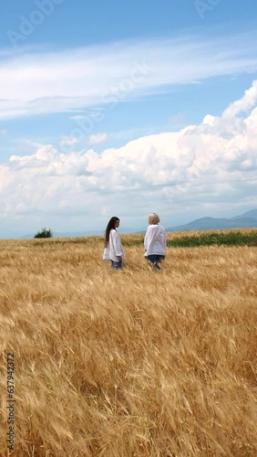 Two girls in white linen shirts and jeans walking in a rye field on a windy day