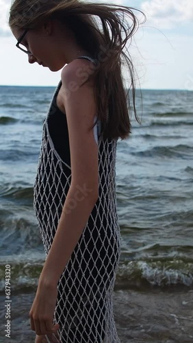 Teenage girl with long brown hair in glasses and net dress strolls through coastal waves