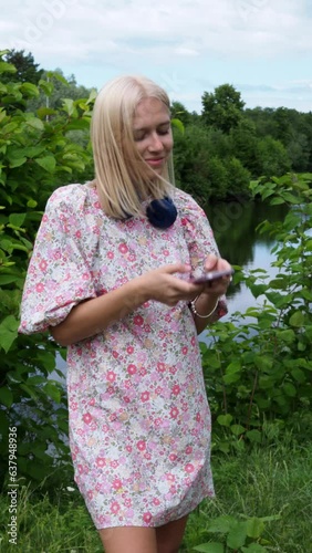 A young blonde in a floral dress stands by the lake and looks at her smartphone