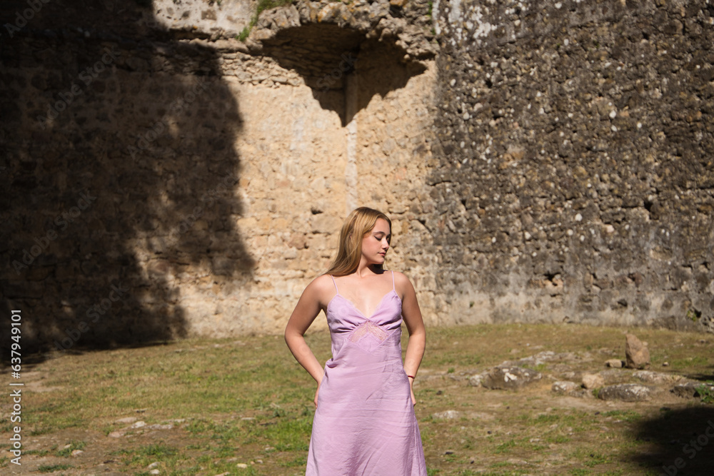 Young beautiful blonde woman walking relaxed in fortress, medieval castle in spain. The woman is wearing a long violet dress. Travel and holiday concept.