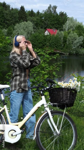 Young woman with bicycle and flowers listens to music in headphones by pond