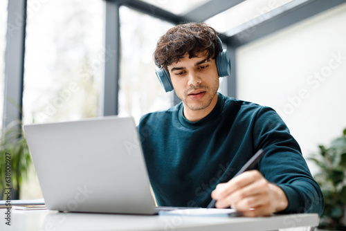 Remote studying. Concentrated european guy in headphones watching video course on laptop and taking notes