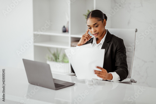 Business paperwork. Black businesswoman reading papers, sitting in front of laptop computer in light modern office photo