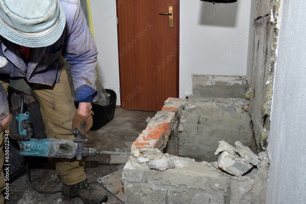 A bricklayer uses a jackhammer to break a wall in a house while building a fireplace