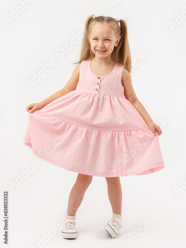 A 5-year-old Caucasian girl with long hair stands on a white background smiling, holding a pink dress with her hands. The legs are crooked.
