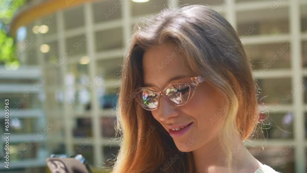 Woman using mobile phone standing in background of cafe in park on summer day. Happy caucasian female person in glasses with cell phone looking device screen browsing internet Woman using mobile phone standing in background of cafe in park on summer day. Happy caucasian female person in glasses with cell phone looking device screen browsing internet