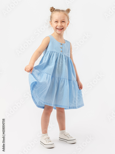 Happiness, joy. A 5-year-old girl with a fashionable hairstyle is standing sideways on a white background, holding a blue summer dress with her hand and laughing. photo