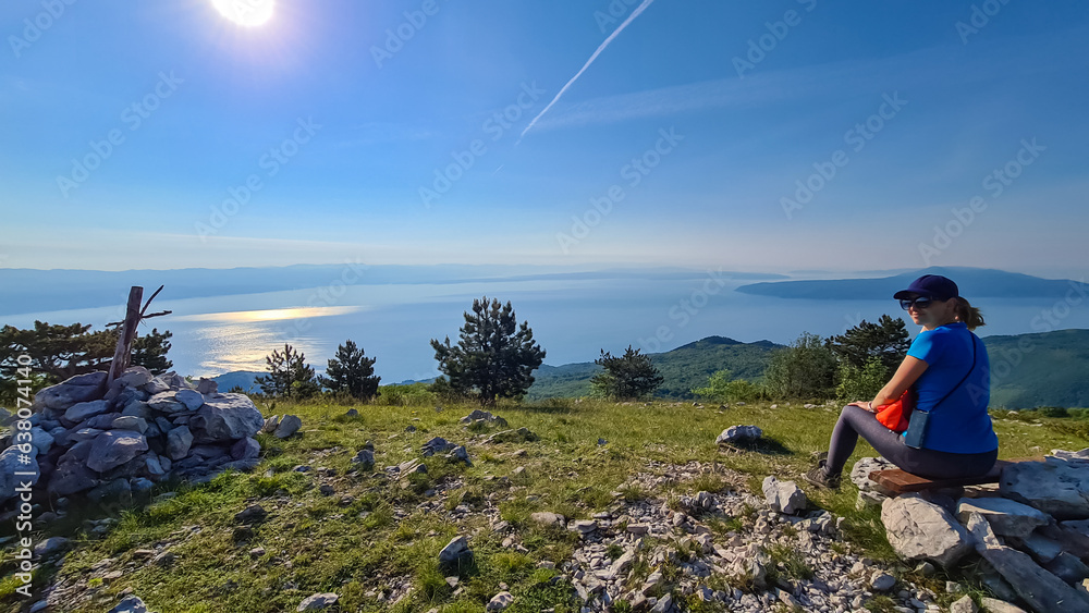 A woman sitting on top of Vojak and enjoying the panoramic view on Mediterranean Sea. The mountain is overgrown with lush green plants. Few islands in the back. Early morning hiking by the sea. Remedy