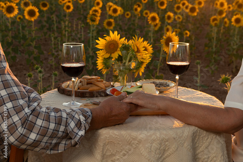hands of senior couple toasting with glasses of wine having dinner al fresco in sunflower fields
