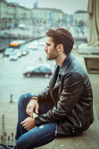 Photo of a stylish man in a leather jacket posing on a ledge outdoors