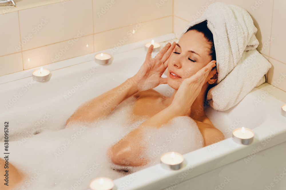 The girl massages the temples of her head, relaxing in a bath with foam and candles. The girl massages the temples of her head, relaxing in a bath with foam and candles.