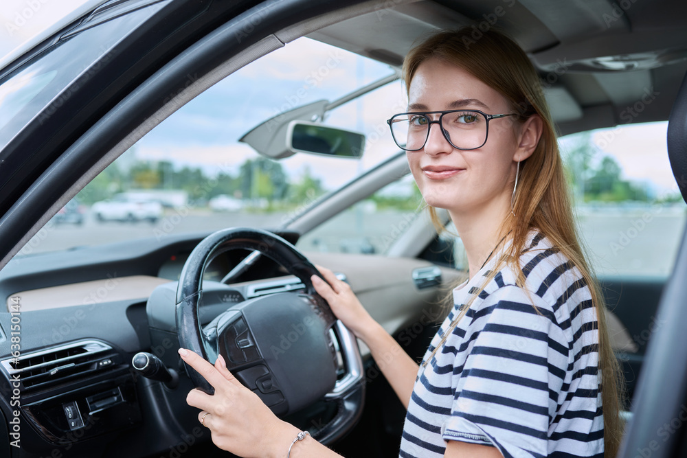 Teenage girl driver in glasses sitting behind wheel of car, looking at camera
