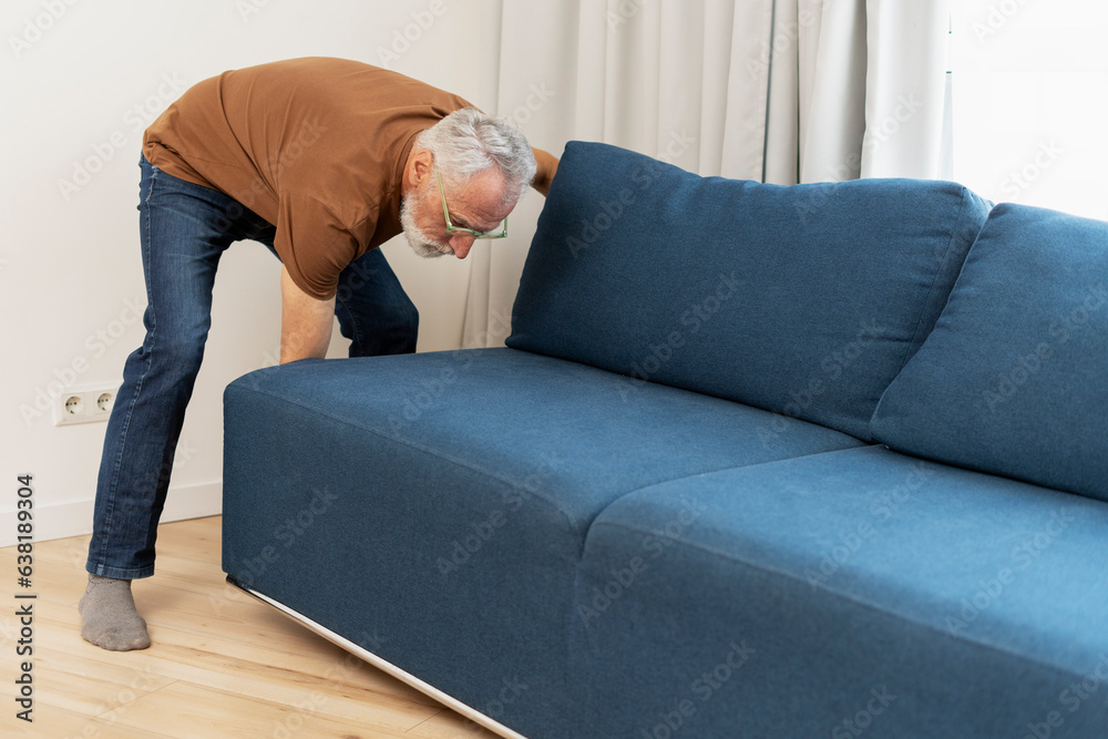 Portrait senior man in glasses moving sofa in apartment. Elderly pensioner in living room