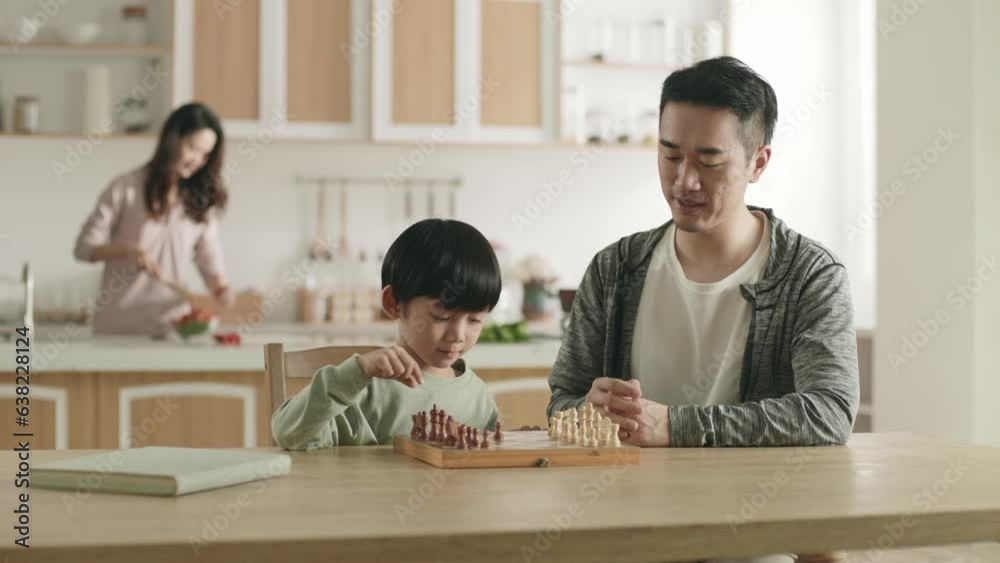 young asian father playing chess with five-year-old son at home while mother preparing meal in family kitchen young asian father playing chess with five-year-old son at home while mother preparing meal in family kitchen