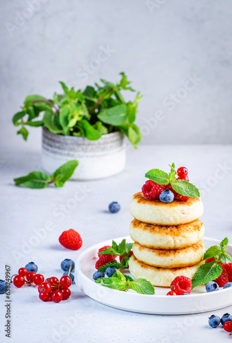 Thick pancakes with cottage cheese with blueberries  raspberries and red currants  decorated with mint on white plate  delicious breakfast. Gray table background