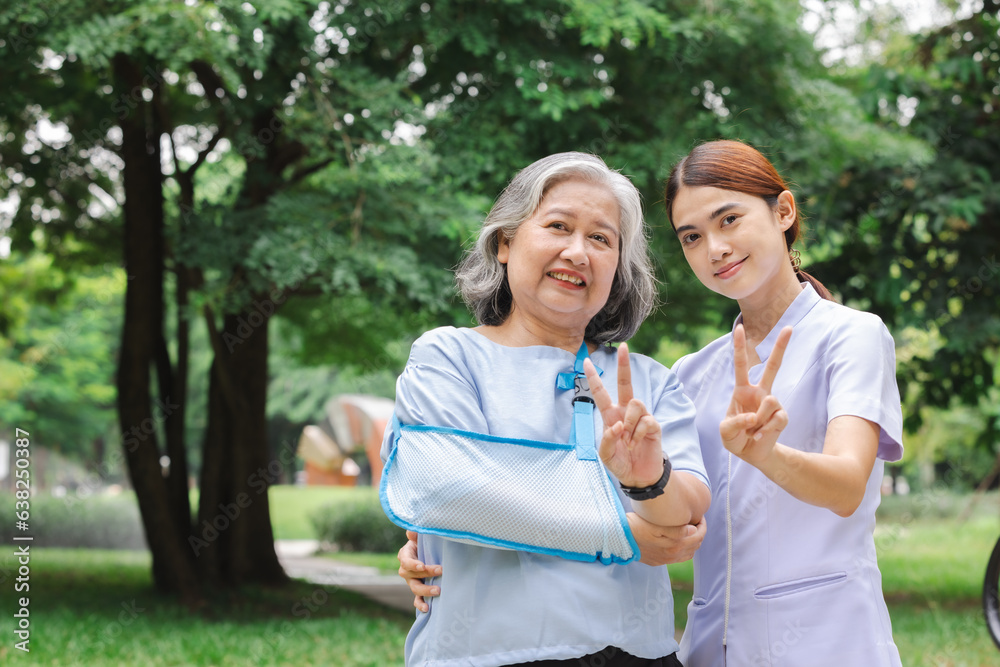 Senior Asian woman wearing patient gown and soft splint due to sore arm showing okay sign with nurse, personal accident concept. Senior Asian woman wearing patient gown and soft splint due to sore arm showing okay sign with nurse, personal accident concept.