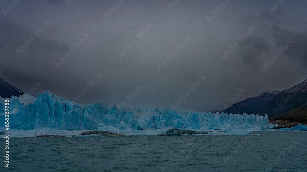 Naklejka premium A wall of blue ice with sharp spikes rises above a turquoise lake. Cloudy. Mountains in the fog. Icebergs float in the water. Perito Moreno glacier. Los Glaciares National Park. El Calafate. Argentina