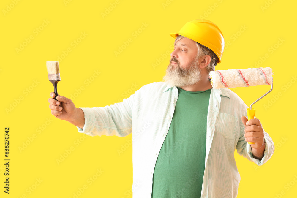 Portrait of senior man in hardhat with paint roller and brush on yellow background. Labor Day celebration